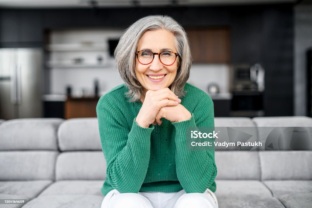 Healthy aged woman sitting on the sofa Healthy aged woman with grey hair with glowing blue eyes, elegant glasses, sitting on the pure comfortable sofa at modern apartment, dressed in green sweater and white pants, put chin on the hands One Woman Only Stock Photo