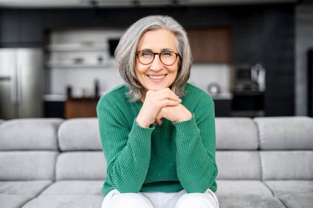 Healthy aged woman sitting on the sofa Healthy aged woman with grey hair with glowing blue eyes, elegant glasses, sitting on the pure comfortable sofa at modern apartment, dressed in green sweater and white pants, put chin on the hands innocence stock pictures, royalty-free photos & images