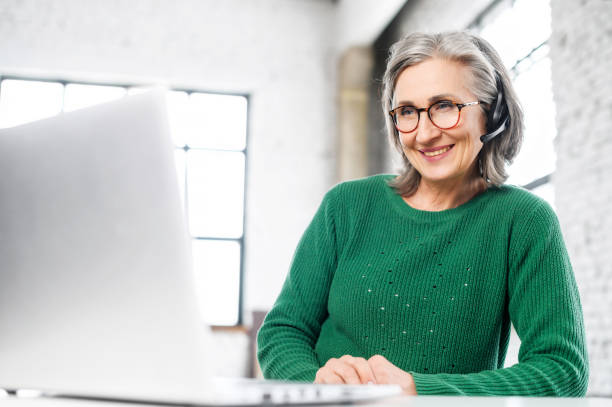 cheerful aged woman with headset sitting at the computer - bluetooth headset women customer imagens e fotografias de stock