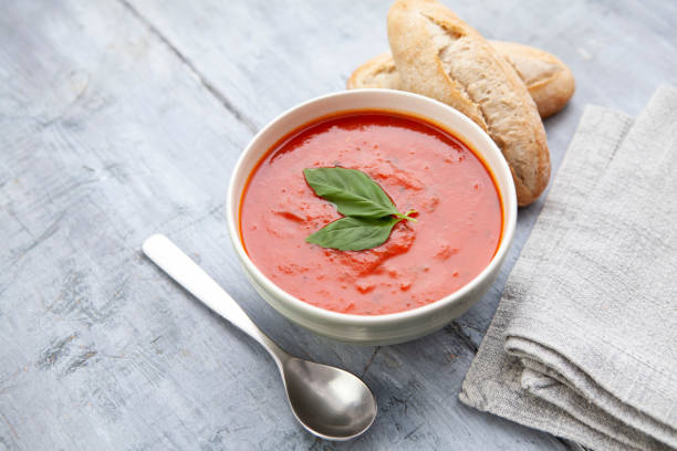 sopa de tomate con hojas de albahaca en un tazón verde con dos panecillos, una cuchara y una servilleta sobre una mesa de madera rústica - sopa de tomate fotografías e imágenes de stock