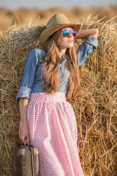 girl in a wheat field wearing hat and sunglasses and holding suitcase