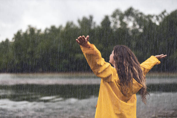 Back view of a carefree woman on rain by the river. Rear view of carefree woman standing in nature with her arms outstretched during rainy day. Copy space. rain stock pictures, royalty-free photos & images