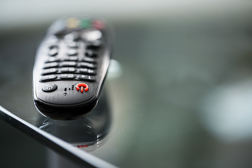 Black TV remote control with red button lying on glass table surface. Home entertainment concept.