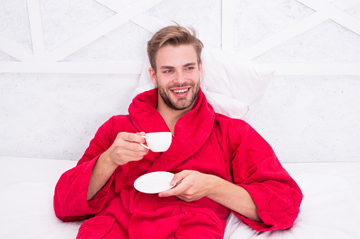 Drinking coffee for breakfast. Happy guy drinking coffee in bed. Bearded man in bathrobe holding drinking cup and smiling. Enjoying drinking tea or other hot beverage after bath spa.