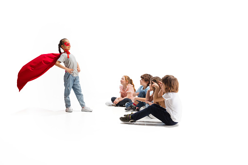 Power. Child pretending to be a superhero with her friends sitting around her. Kids excited, inspired by their strong, brave friend in red coat isolated on white background. Dreams, emotions concept.