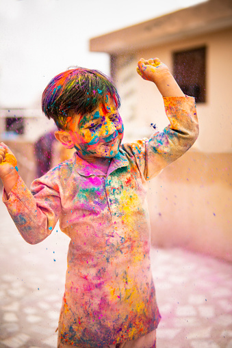 Outdoors image of an Asian/ Indian happy little boy celebrating Holi festival with color powder on rooftop at day time.