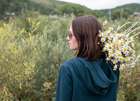 Rear view of young woman in sunglasses holding bouquet of daisies on her shoulder