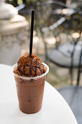 a glass of iced dark chocolate, one topped with chocolate ice cream, glass placed on white table with cafe environment.