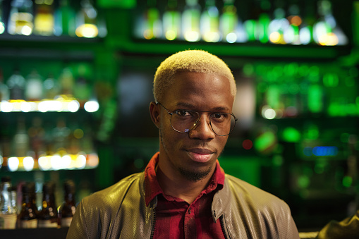 Young African man in smart casualwear and eyeglasses looking at you while standing in front of camera against bar with alcoholic drinks