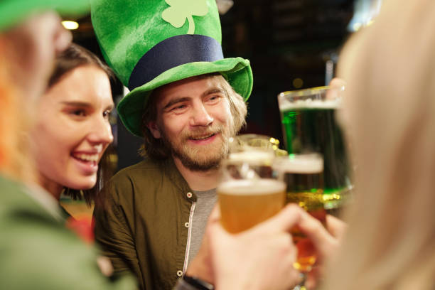 felice giovane barbuto in tradizionale cappello verde irlandese tostato con i suoi amici - irish culture beer drinking pub foto e immagini stock