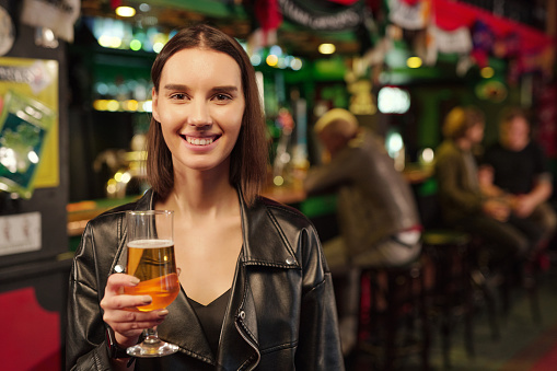 Young cheerful brunette woman looking at you while toasting with glass of beer in front of camera against guys sitting by bar counter in pub