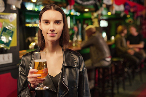 Portrait of young woman holding glass of beer and looking at camera standing in the pub
