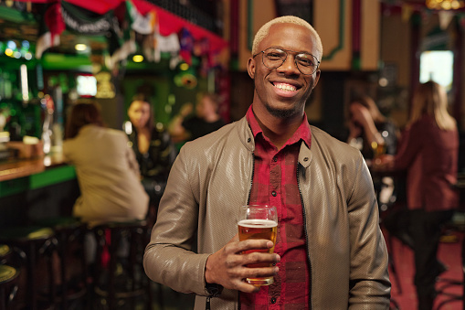 Portrait of African young man holding glass of beer and smiling at camera while standing in the bar