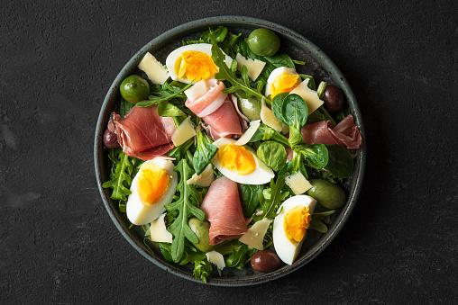 Salad with prosciutto, olives, cheese, eggs and arugula in a plate on black background. Italian food snacks. Top view. Healthy diet food