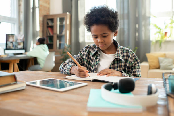 istock Cute elementary African schoolboy with pencil making notes in copybook 1304252794