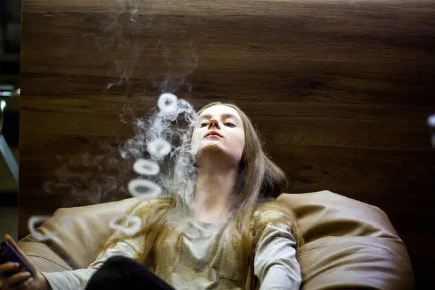Vaping teenager. Young pretty white caucasian teenage girl with problem skin smoking an electronic cigarette indoors. Deadly bad habit. Vape activity.