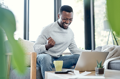 Shot of a young man celebrating while going over paperwork and using a laptop at home