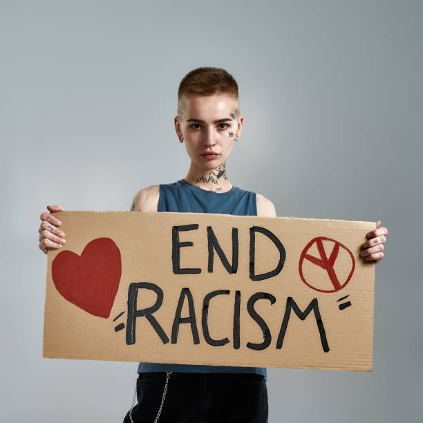 Attractive tattooed young caucasian woman with short hair looking at camera, holding cardboard End Racism banner in front of her, posing isolated over gray background Attractive tattooed young caucasian woman with short hair looking at camera, holding cardboard End Racism banner in front of her, posing isolated over gray background. Social issues, protest concept ending racism stock pictures, royalty-free photos & images