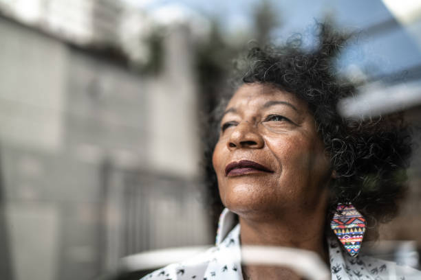 femme aîné regardant par la fenêtre à la maison - regarder par la fenêtre photos et images de collection