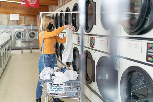 An attractive and stylish caucasian millennial woman stands next to a row of washing machines and dryers and folds her clean laundry in a laundromat. She is wearing a protective face mask because of the coronavirus and business regulations.