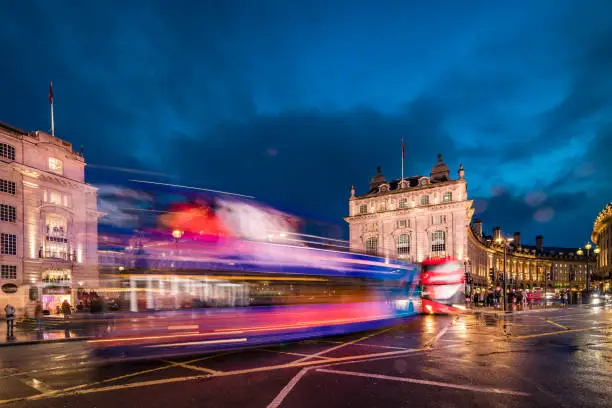 Long exposure photo with evening illumination and reflections from the wet roads at Piccadilly Circus in London city during blue hour on a rainy English evening. Crowd and traffic with motion blur due to the exposure time with unrecognisable accidental commuters and tourist. Image has copy space and it is ideal for background. Shot on Canon EOS R full frame system.
