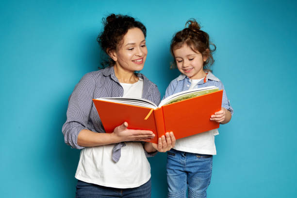 woman and girl equally dressed hold a book in their hands, smiling tenderly, looking at it. blue background with copy space - child looking blank offspring imagens e fotografias de stock