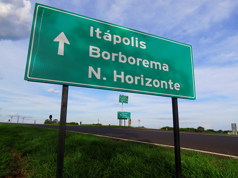 Metal plate giving the direction of three cities (names displayed in portuguese) with a big white arrow placed at the sideway of a countryside route in Brazil in vibrant colors