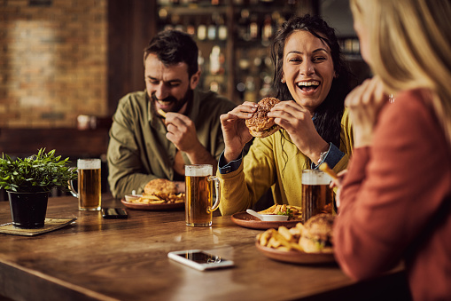 Group of happy friends talking and having fun while eating hamburgers and drinking beer in a pub. Focus is on woman in the middle.