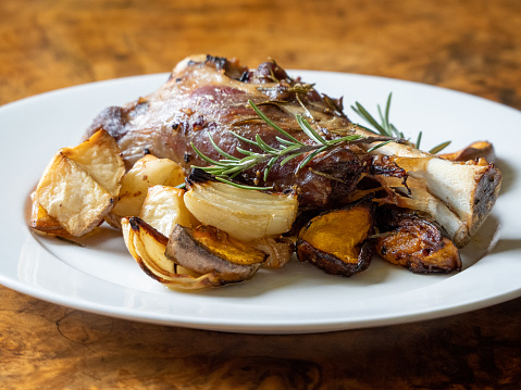 Closeup photo of a home cooked roasted lamb shank with fresh rosemary and roasted potato, sweet potato and pumpkin on a round white ceramic plate.