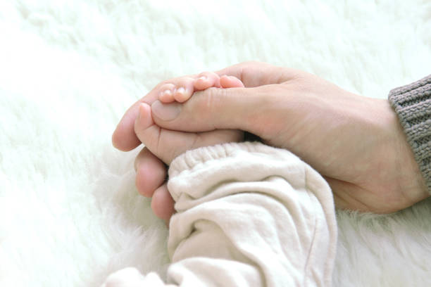 The image captures a tender moment between an adult and a baby, focusing on their hands. The adult's hand, weathered and strong, gently holds the tiny, delicate hand of the baby. The contrast between the rough texture and larger size of the adult's hand and the softness and fragility of the baby's fingers highlights the themes of care, protection, and love. The baby is dressed in a soft, knitted white garment, adding a sense of warmth and purity to the scene. The background is softly blurred, ensuring that the viewers attention is drawn entirely to the touching connection between the two hands, symbolizing a bond that is both nurturing and profound.