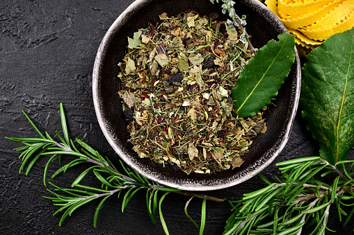 Fresh and dry aromatic condiments in a bowl with green rosemary on a black rustic background. Dry green herbs for preparing the tea