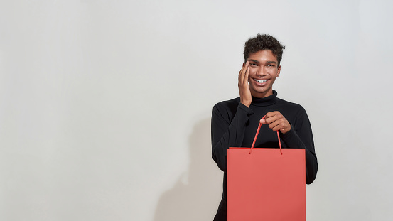 Surprised young african american guy holding shopping packet and looking at camera while standing on light background, widescreen. Discount and sale concept