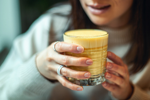 Young caucasian woman in warm sweater is smiling and holding a glass of hot turmeric milk on table of coffee shop. Enjoyment of drinking hot beverage near window on weekend. Copy space