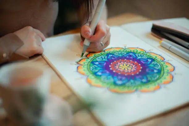 Photo of Young woman colouring mandala with markers and white rosary on table with cup of coffee at home