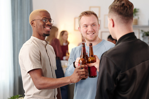 Happy young intercultural men clinking with bottles of beer and looking at one another with toothy smiles while cheering up at home party