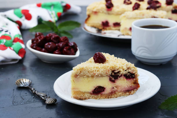 Cottage cake with cherries located on a plate on dark background, piece of cake and a cup of coffee in the foreground