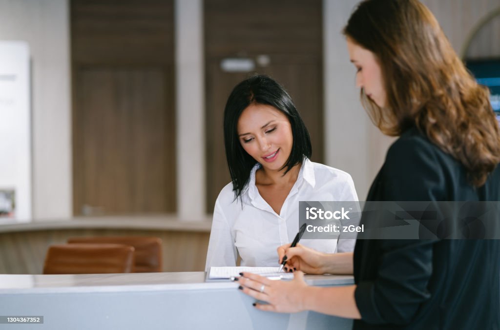 Portrait of friendly young woman reception standing behind the reception at desk of hotel with customer visitor check-in at hotel. Hotel Reception Stock Photo