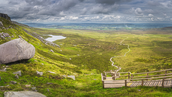 Large panorama with view on steep stairs of wooden boardwalk leading to Cuilcagh Mountain peak with lake and valley or plane below, Northern Ireland
