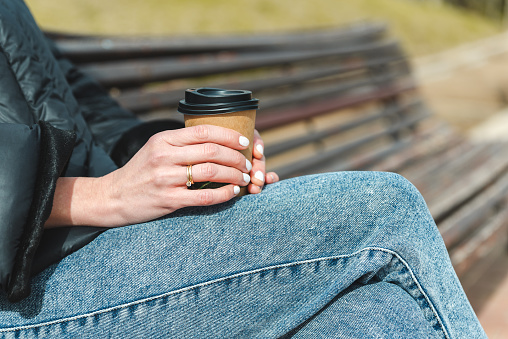 close-up take of the hands of a white woman holding a disposable paper cup of coffee. She wears blue jeans, black coat and a ring on her fingers. She is sitting on a wooden bench at an urban green park