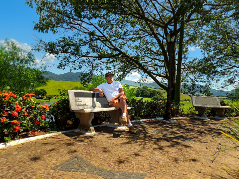 Beautiful colorful image of an old man wearing beret and glasses sitting alone on park bench surrounded by nature in a small town in the countryside of brazil called Joanopolis