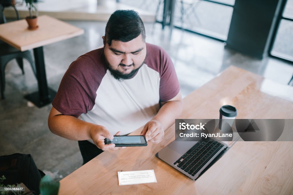 Young Adult Doing Remote Capture Deposit With Check A Hispanic man in his 20's works on his computer in a public cafe, enjoying a latte in a paper cup.  He takes a photo of a paycheck with his smartphone for mobile deposit to his bank checking account. Bank Deposit Slip Stock Photo
