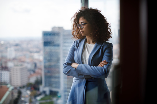 Young thoughtful businesswoman wearing a suit standing in her office and looking through window