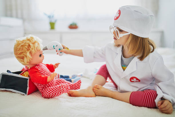 Adorable toddler girl in white coat playing doctor and giving medical care to her doll Adorable toddler girl in white coat playing doctor and giving medical care to her doll. Children and role games. Covid-19 pandemic concept girl playing with doll stock pictures, royalty-free photos & images