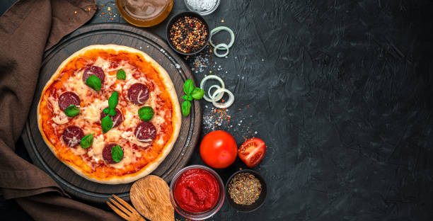 istock Culinary panorama with pizza with salami and cheese and ingredients on a black background 1304395740
