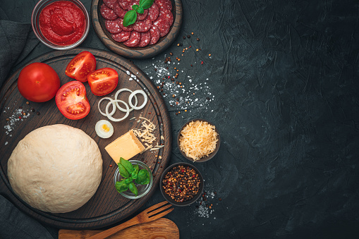 Dough, salami, tomatoes, cheese, basil and spices on a black background. Ingredients for pizza. Cooking concept.