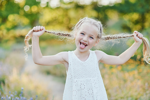 Funny girl holding hair plaits. Small cute girl with long blonde hair showing tongue outdoor. Kid expressing emotions. April fool