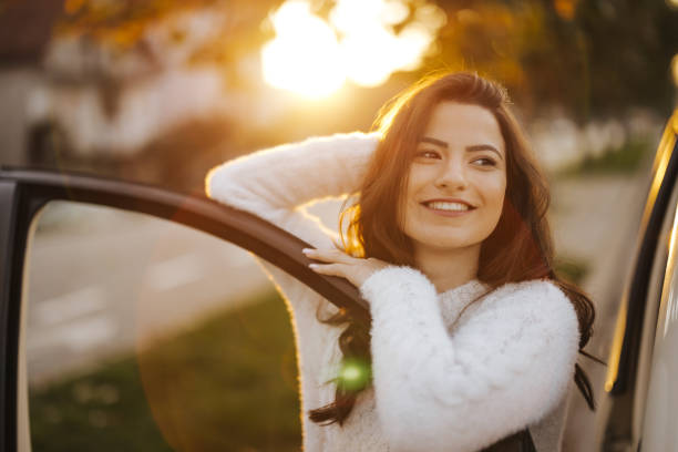 Beautiful young smiling woman with long brown hair in white sweater standing by her car and enjoying autumn sunset in the city