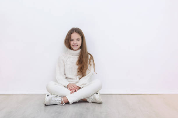 1 white girl 10 years old in a white sweater, jeans and sneakers sits on the floor against a white wall and smiles, teenager - 10 11 years child human face female imagens e fotografias de stock