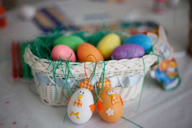 Color image depicting an overhead view of an Easter-themed flat lay on a white wooden background. A basket of red painted eggs is in the foreground, with the white wooden surface defocused beyond.