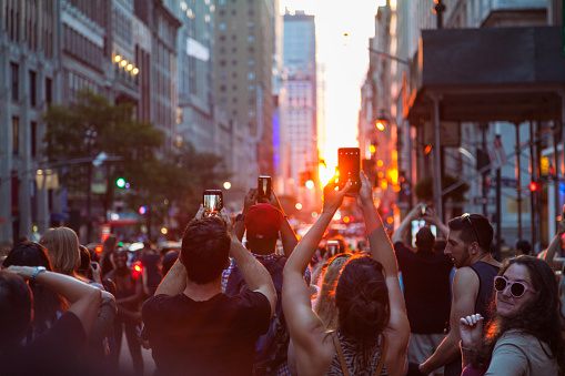 View of the street and city life of Manhattan in the Twilight hours as the sun sets over in New York City. Manhattanhenge, also called the Manhattan Solstice, is an event during which the setting sun or the rising sun is aligned with the east–west streets of the main street grid of Manhattan. People are holding telephones over their heads. Evening. July 13, 2018. NYC. Midtown Manhattan. New York. USA